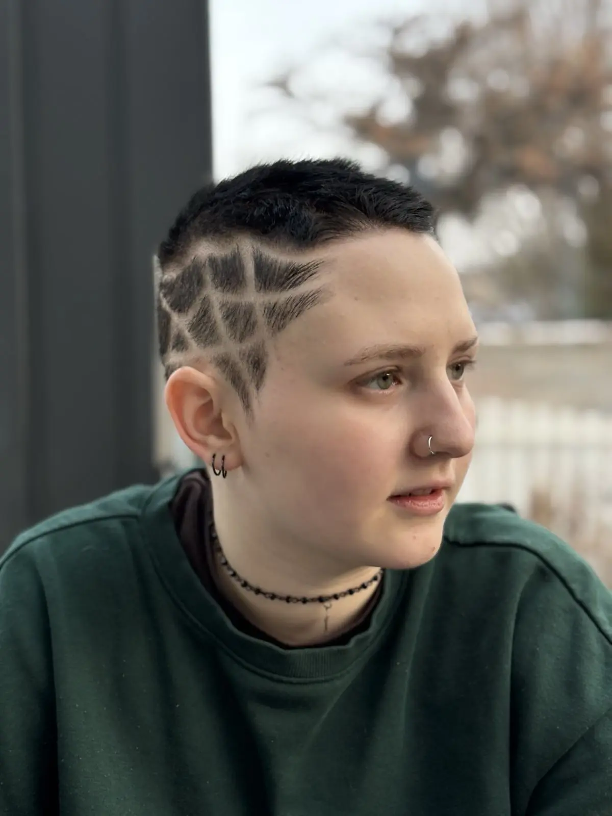 Porträtfoto des 17-jährigen Moony. Kurze, schwarz gefärbte Haare mit einem einrasierten Muster auf der Seite des Kopfes. 2 Piercings im Ohr, eines in der Nase. Grau-grüne Augen. Weiße Hautfarbe. Dunkelgrüner Sweater.