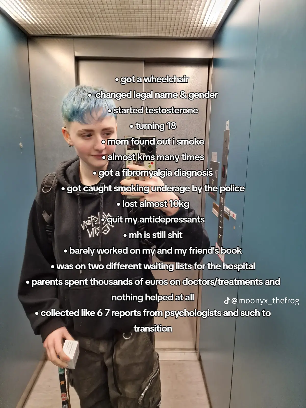 Foto von Moony, der in einem Aufzug steht und ein Selfie macht. Kurze blaue Haare, helle Haut, schwarzer Hoodie, olivgrüne Hose. In der rechten Hand hält er eine kleine weiße Medikamentenschachtel. Darüber sind einige Textzeilen eingeblendet: • got a wheelchair • changed legal name & gender • started testosterone • turning 18 • mom found out i smoke • almost kms many times • got a fibromyalgia diagnosis • got caught smoking underage by the police • lost almost 10kg • quit my antidepressants • mh is still shit • barely worked on my and my friend's book • was on two different waiting lists for the hospital • parents spent thousands of euros on doctors/treatments and nothing helped at all • collected like 6 7 reports from psychologists and such to transition